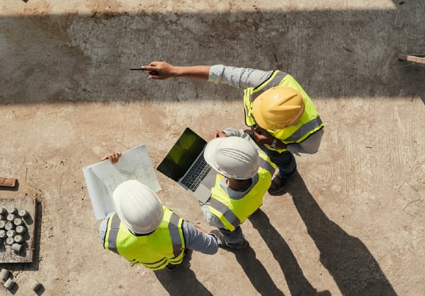 Contractors reviewing plans and laptop on an active construction site from overhead view.
