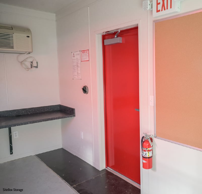Interior of mobile office building with work counter, exit door, bulletin board, and fire extinguisher.